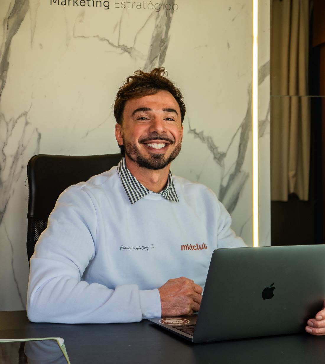 Hagner Saenger sorrindo em sua mesa no estúdio da Praevia, com o logo da agência na parede de mármore