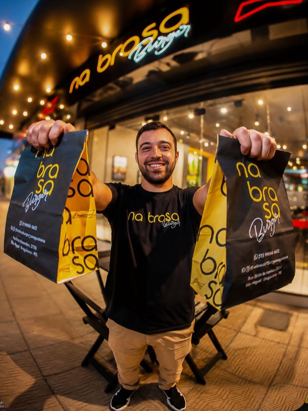 Atendente do Na Brasa Burger segurando duas sacolas de delivery em frente à loja